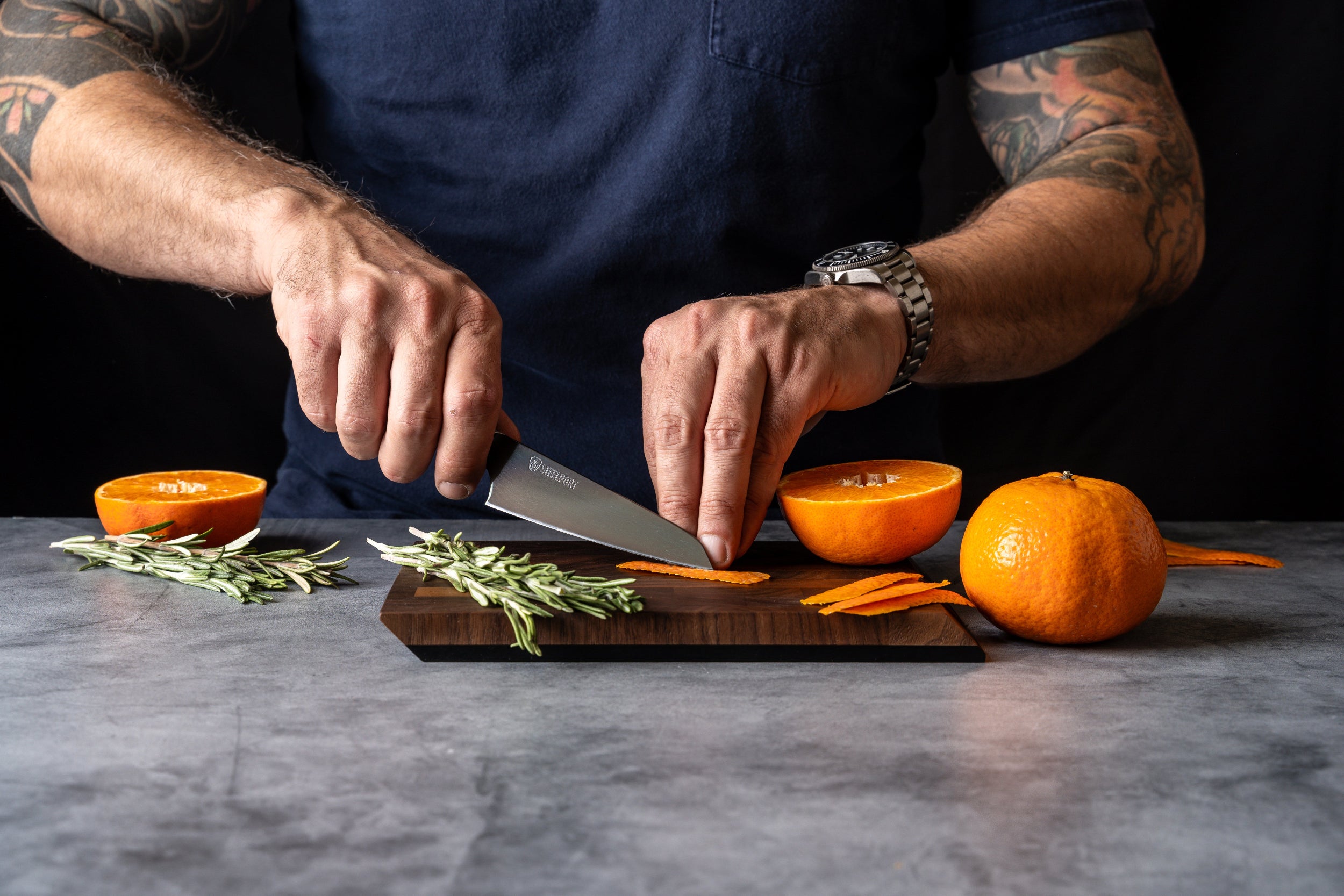 Person cutting vegetables on a SteelCore™ cutting board with oranges and herbs on a dark surface