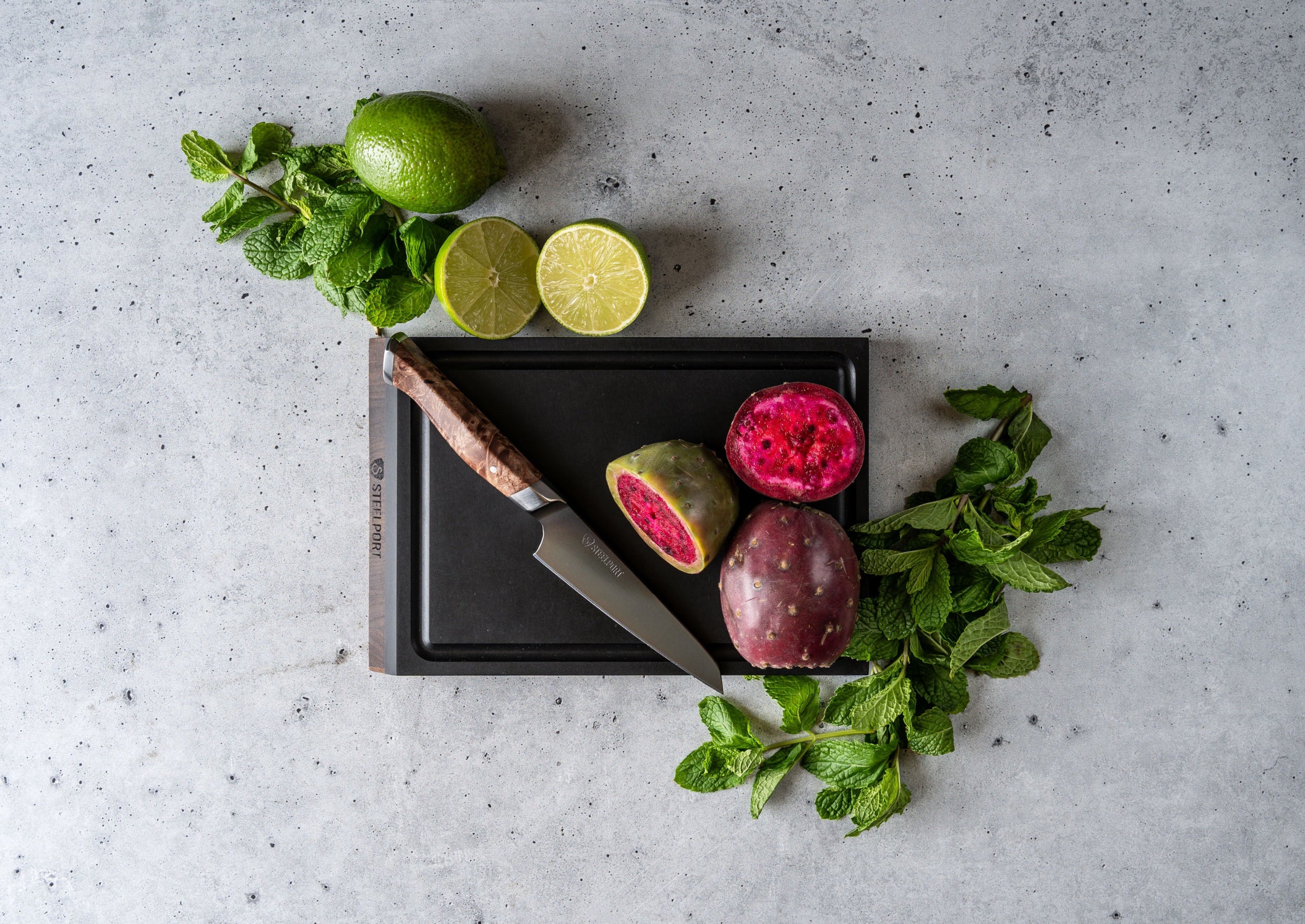 Sliced pomegranate, limes, and a knife on a SteelCore™ cutting board with mint leaves on a gray surface.
